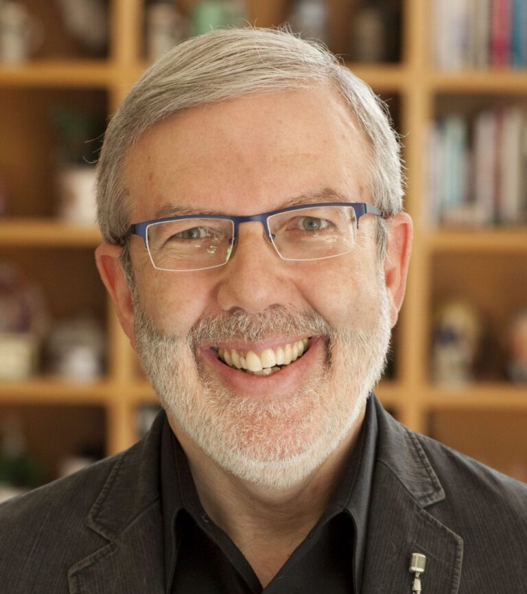 Leonard Maltin smiling, wearing glasses and a dark jacket, with a bookshelf background.