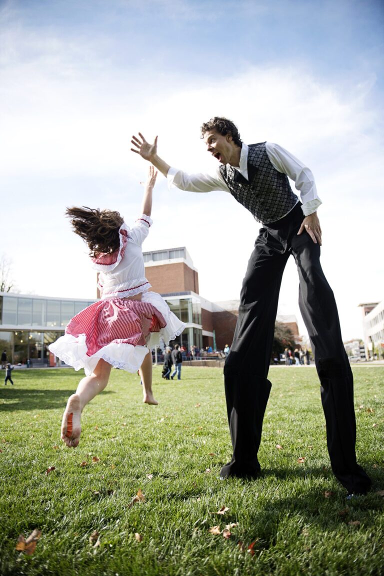 Stilt walker and girl in costume interacting on grassy lawn.