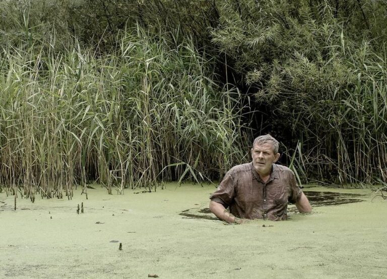 Man stands chest-deep in a green, algae-covered swamp, surrounded by tall reeds and foliage.