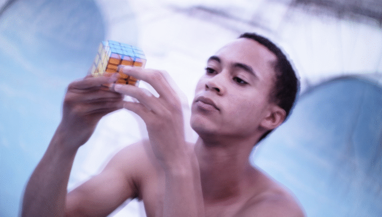 Man solving a Rubik's Cube, focused and shirtless, against a blurred background.