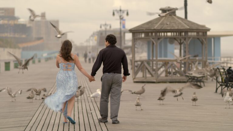 Couple walks hand-in-hand on a boardwalk with seagulls around them.