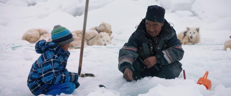 Inuit man and child ice fishing with sled dogs nearby.