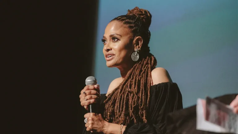 Ava DuVernay speaking at an event, wearing a black dress and locs.