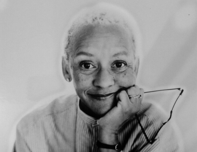 Portrait of Nikki Giovanni, smiling with glasses in hand.