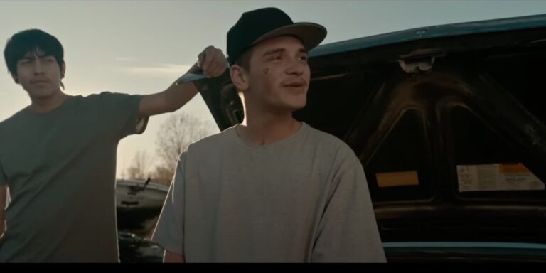 Two young men in 'War Pony' stand by a car with the hood open, one holding a knife.