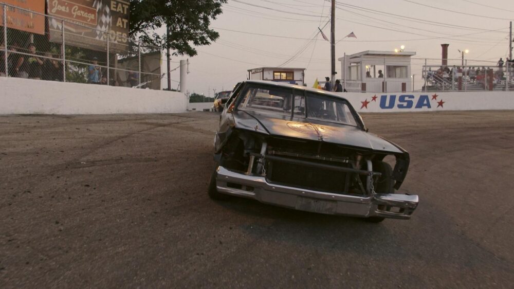 Demolition derby car on track, USA banner in background. "Joe's Garage: Eat at Joe's" sign visible.