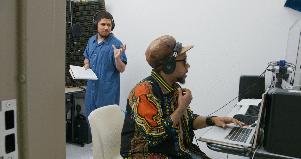 Two men recording music in a studio, one in a blue jumpsuit, the other at a laptop.