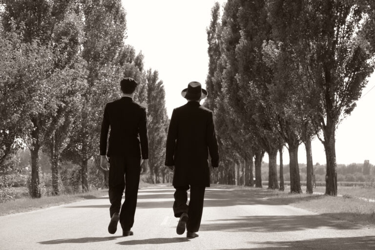 Two men in suits walking down a tree-lined road.