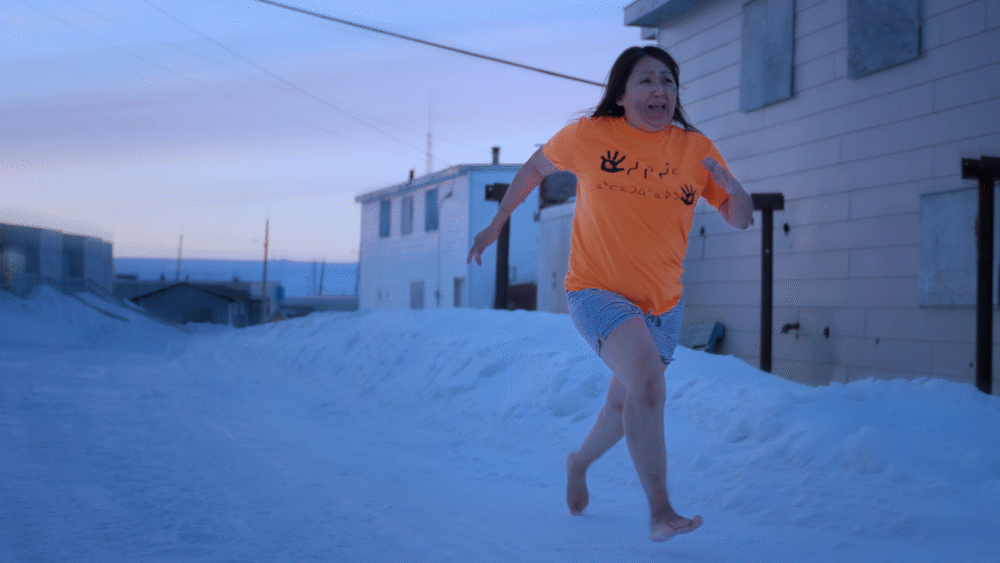 Woman runs barefoot on snow in Tautuktavuk, wearing orange shirt.