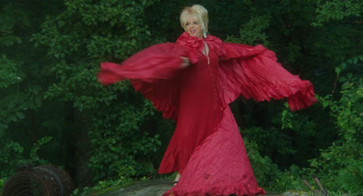 Drag queen in a red gown with flowing sleeves in a garden setting.