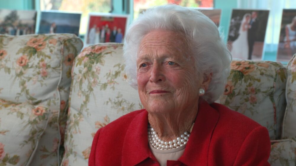 Barbara Bush, former First Lady, wearing a red jacket and pearl necklace, smiling softly.