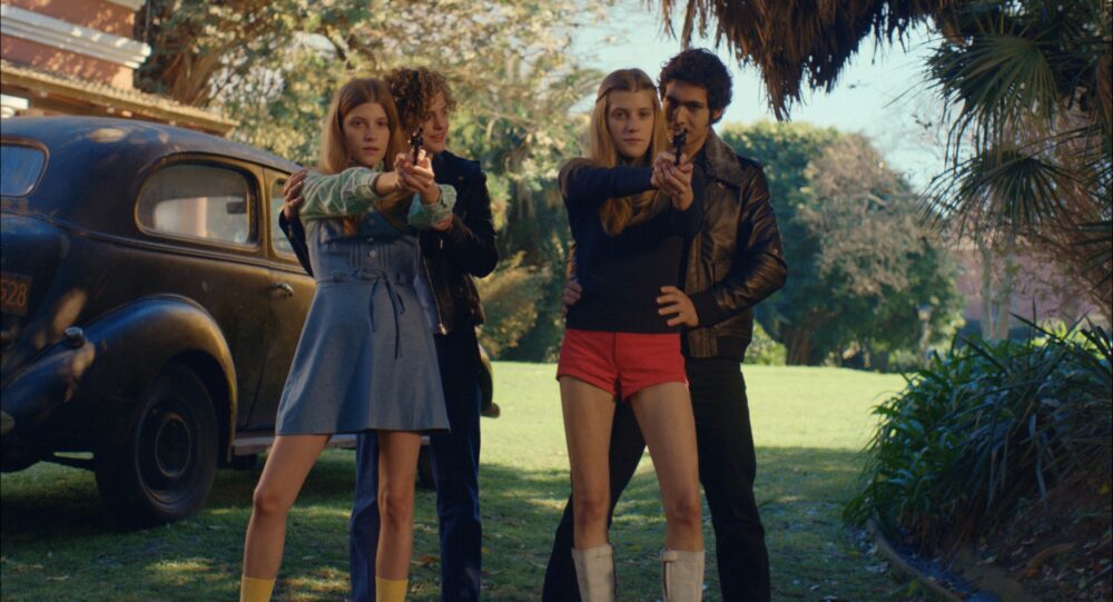 El Angel movie scene: Two couples pointing guns, vintage car in background.