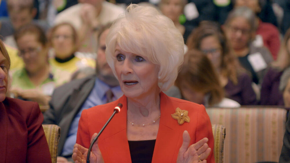 Woman speaking at a hearing, wearing an orange jacket and pearl necklace.