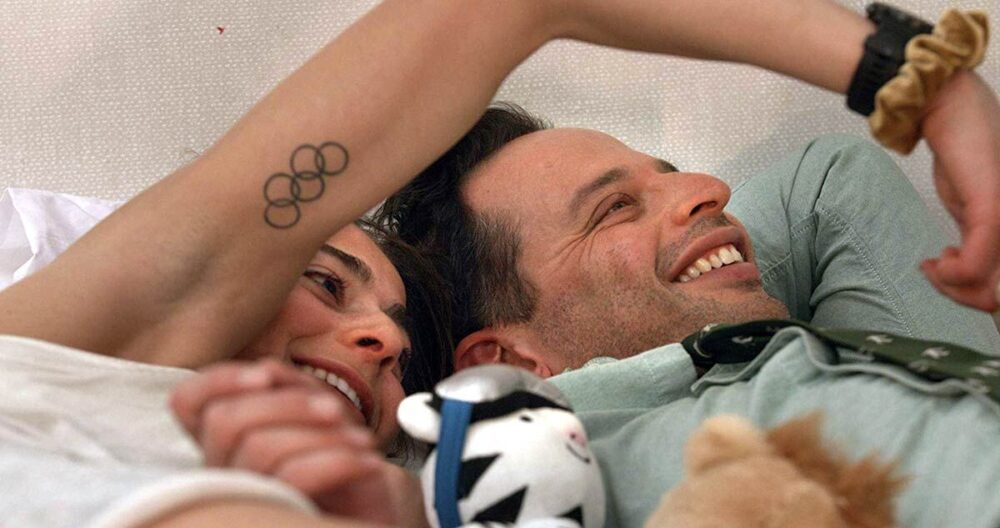 Couple smiling in bed, woman's arm shows Olympic rings tattoo.