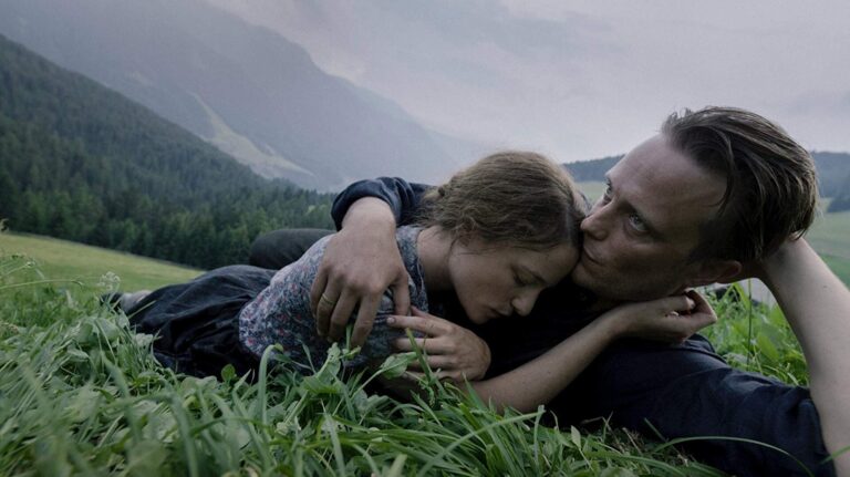 Couple embracing in a field, mountains in the background, from the film A Hidden Life.