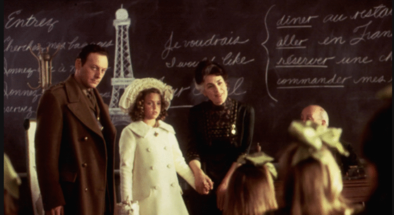 Scene from "A Little Princess" with Shirley Henderson, Liesel Matthews, and Liam Cunningham in a classroom with French phrases on the chalkboard.