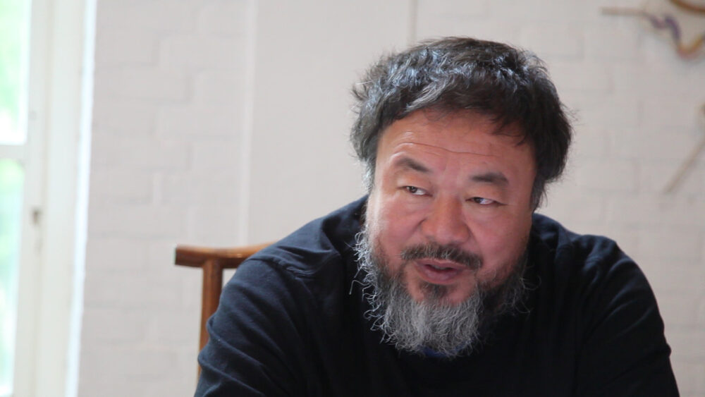 Ai Weiwei portrait, face forward, with salt and pepper beard, wearing a black shirt.