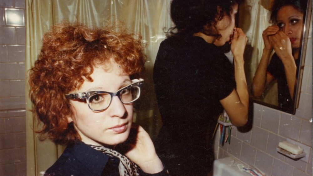 Two women in a bathroom, one with curly red hair and glasses.