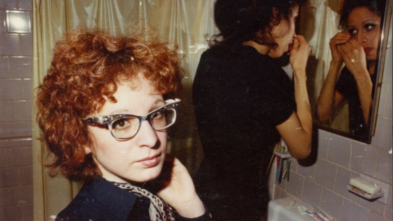 Two women in a bathroom, one with curly red hair and glasses.