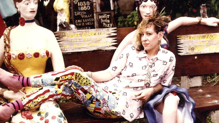 Allee Willis sitting on a bench with folk art statues. "Dentist Painless Pelky" sign visible.