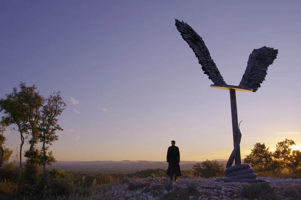 Anselm Kiefer sculpture with wings against a sunset sky, overlooking a valley landscape.