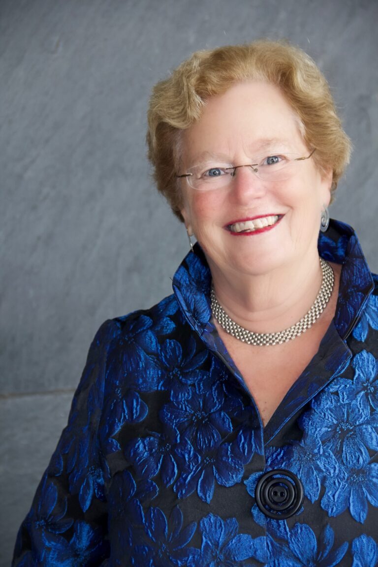 Abby Ginzberg, smiling woman with short blonde hair wearing a blue floral jacket and silver necklace.