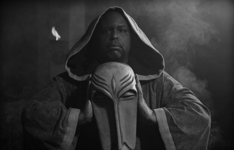 Man in hooded robe holding a mask, reminiscent of The Acolyte's mysterious aesthetic.