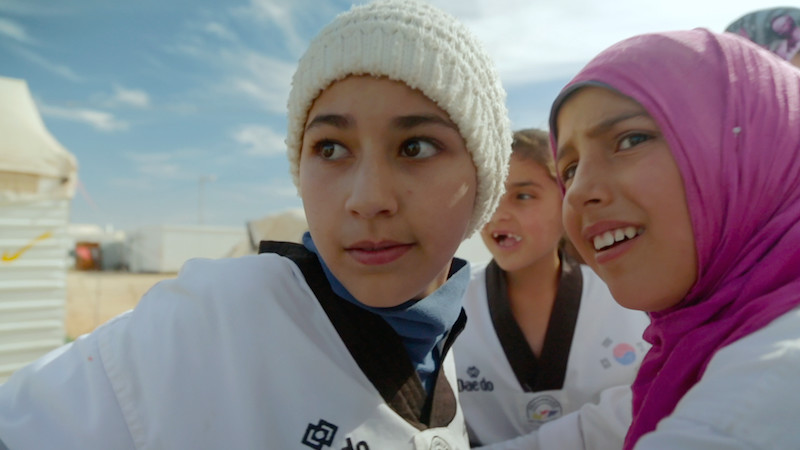 Young girls in taekwondo uniforms, one wearing a hijab and another a beanie, looking off-camera.