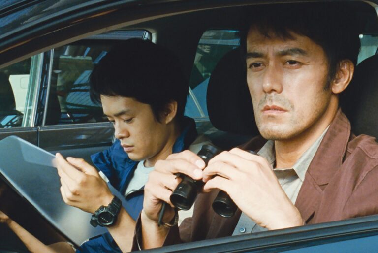 Two men in a car, one with binoculars, the other with a folder.