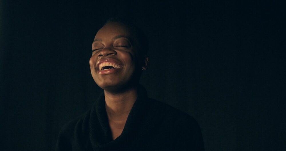 Black woman laughing with eyes closed against a dark background.