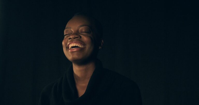 Black woman laughing with eyes closed against a dark background.