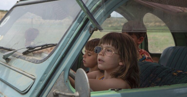Children in a vintage blue car, one wearing sunglasses and pretending to drive in Alcarràs.