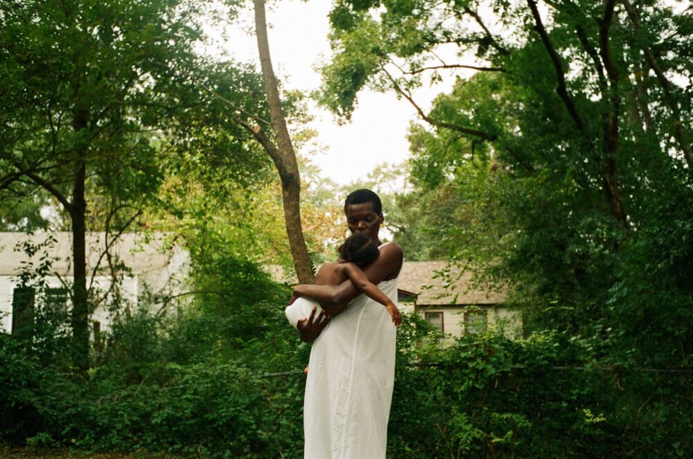 Woman in white dress holding child in lush green outdoor setting. Serene, natural light.