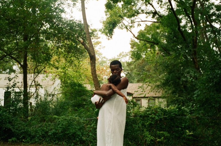 Woman in white dress holding child in lush green outdoor setting. Serene, natural light.