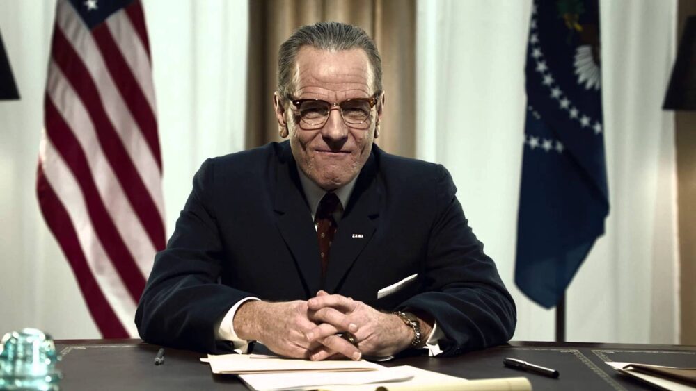 Bryan Cranston as LBJ in "All the Way," sitting at a desk with US and Presidential flags.