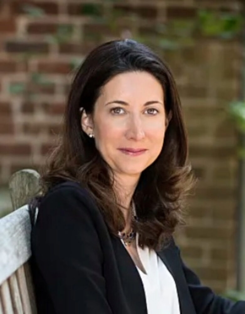 Amanda Cowen portrait. Woman with dark hair in business attire sitting on a bench.