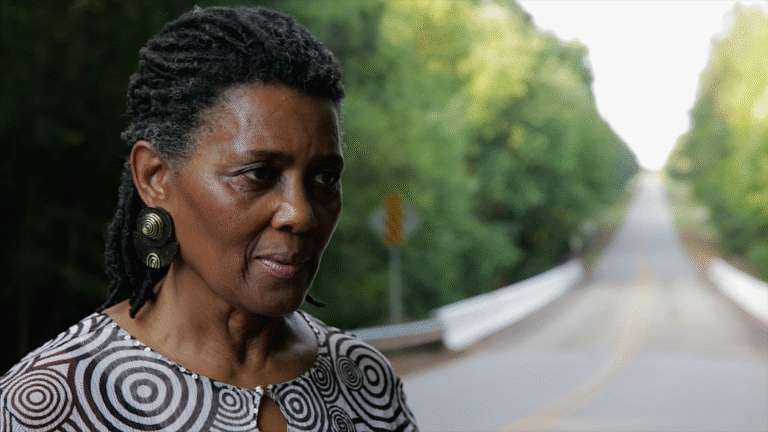 Portrait of a woman with locs standing near a bridge in "An Outrage.