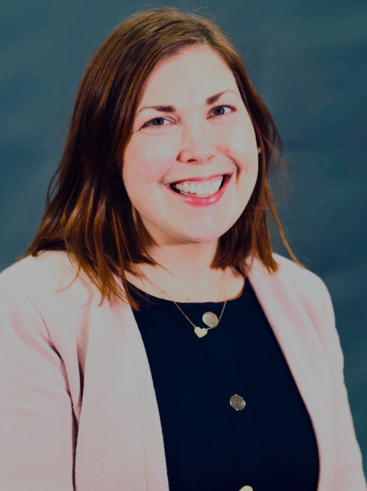 Andrea Goodman smiles, wearing a pink blazer and a necklace with heart pendants.