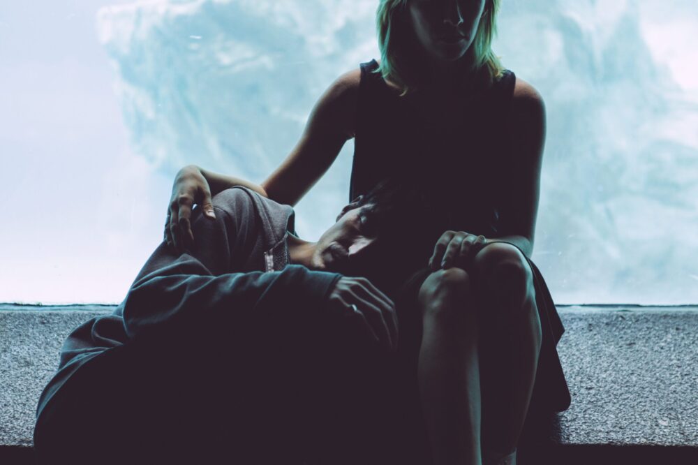 Person resting head on another's lap, sitting against a blue backdrop.