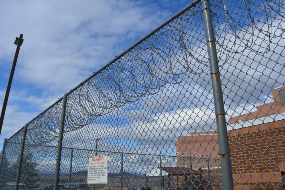 Chain-link fence topped with razor wire, brick building in background. Warning sign: "No Trespassing.