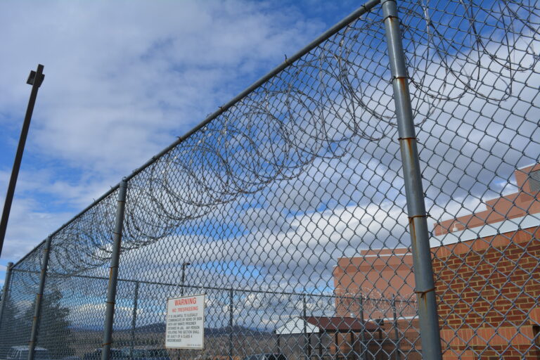 Chain-link fence topped with razor wire, brick building in background. Warning sign: "No Trespassing.