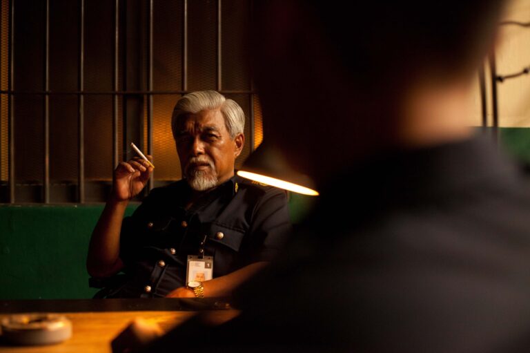 Man with gray hair and beard, in uniform, smoking in front of jail bars.