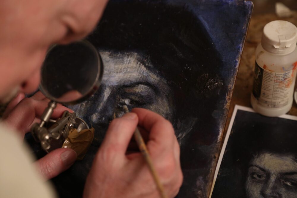 Artist restoring a painting of a face with a magnifying glass.