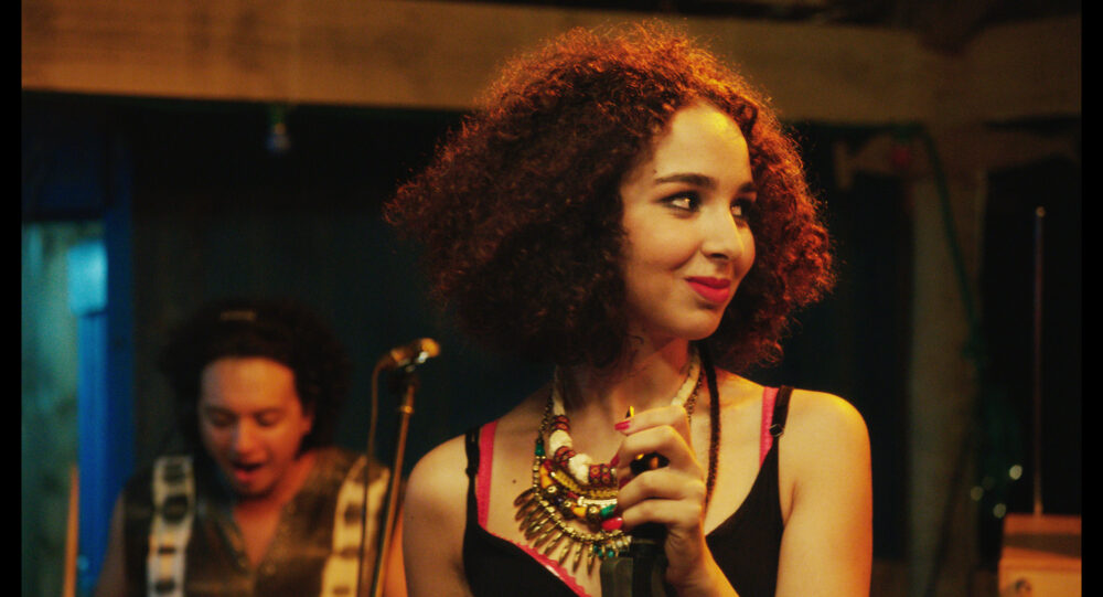Singer with curly hair and colorful necklace performing on stage.