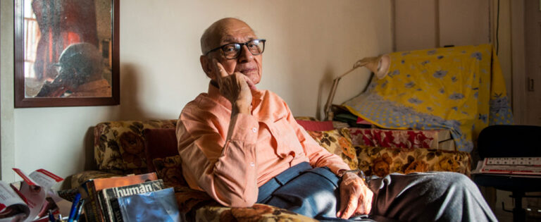 Elderly man with glasses sitting on a couch, appearing thoughtful.