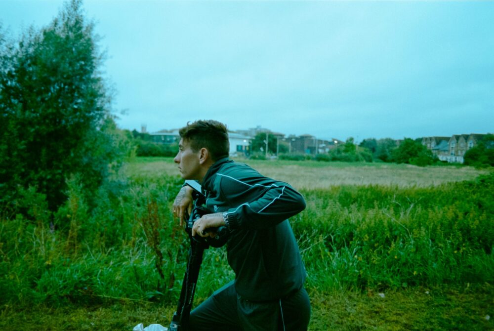 Man in tracksuit with scooter looks out over grassy field.