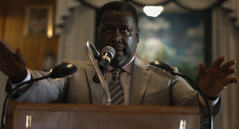 Actor Wendell Pierce speaking at a podium in a tan suit with his hands raised.