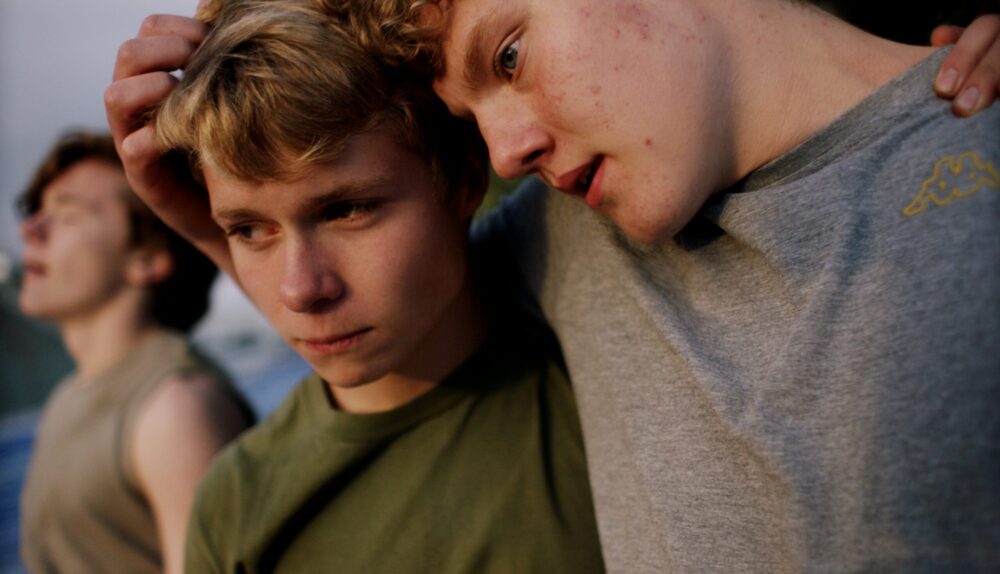 Two young men embracing, one with hand on the other's head. A third man is blurred in background.