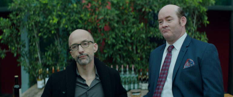 Two men in suits stand outdoors near bottles.