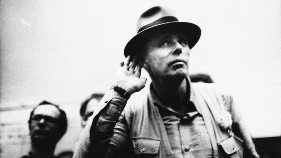 Black and white photo of artist Joseph Beuys wearing a fedora, listening intently.